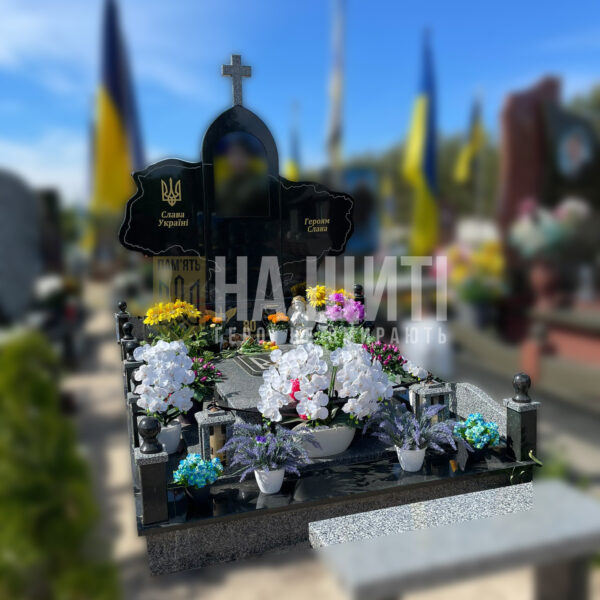 Чорний гранітний пам'ятник Герою у формі карти України з хрестом та квітковим оформленням.
